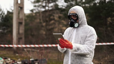 Man in bio-hazard suit and gas mask takes notes in his tablet standing on the polluted land
