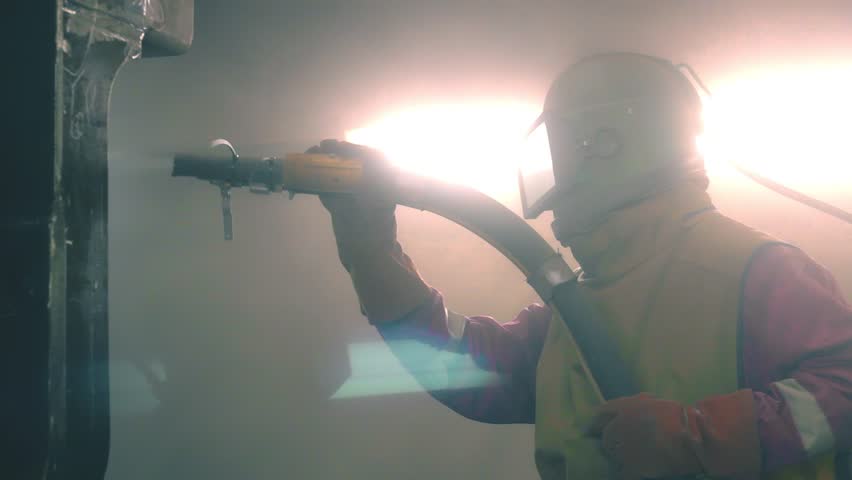 Industrial worker cleans metal surface by sandblaster gun