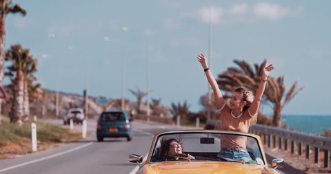 Young women having fun driving convertible vintage car on exotic island palm tree seaside road