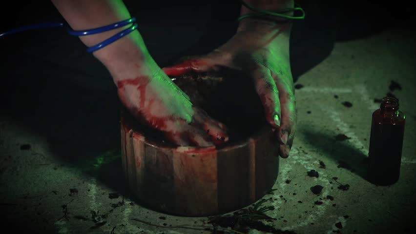 A witch mixes bloody ingredients to make a potion in a wood bowl.