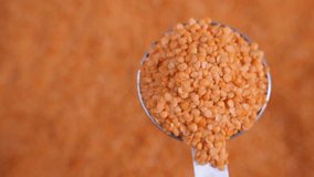 Slow motion bulk dry red lentils tipping a portion of legumes, with a metal kitchen utensil onto an orange surface landscape background with a pile of grains, wholesale of larder store food 