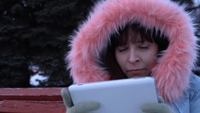 Portrait of a young happy woman uses a digital tablet and sitting on a bench in a city park next to a Christmas tree on a snowy winter day.