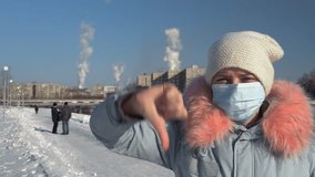 Blogger wearing a protective mask and a gray parka records video on the phone and stands on a background of pipes of a metallurgical plant on a winter day. A young woman is showing a thumbs-down.
