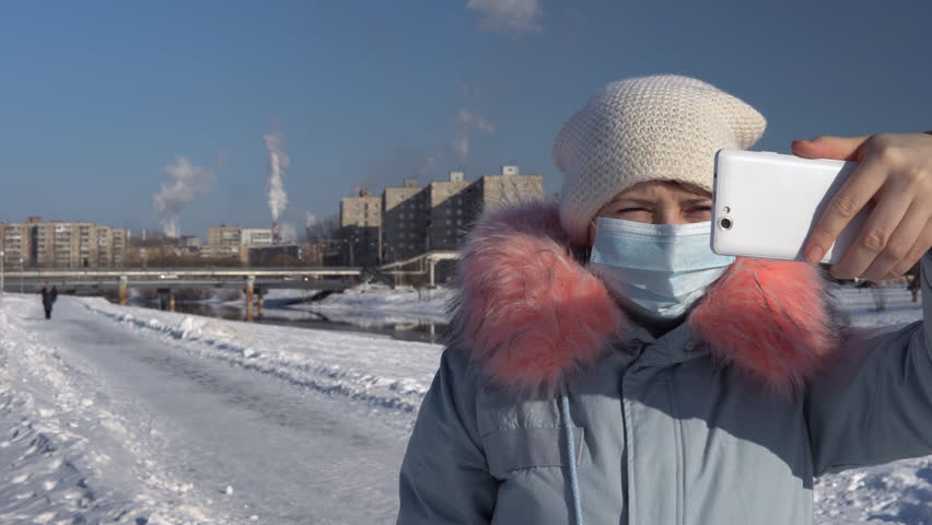 Blogger wearing a protective mask and a gray parka records video on the phone and stands on a background of pipes of a metallurgical plant on a winter day. A young woman is showing a thumbs-down. | Shutterstock HD Video #1010403143