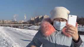 A young blogger wearing a protective mask and a gray parka records video on the phone and stands on a background of pipes of a metallurgical plant on a winter day. A woman waving her hand, greeting.