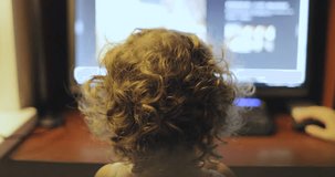 little girl with curly hair sitting near computer monitor rear view