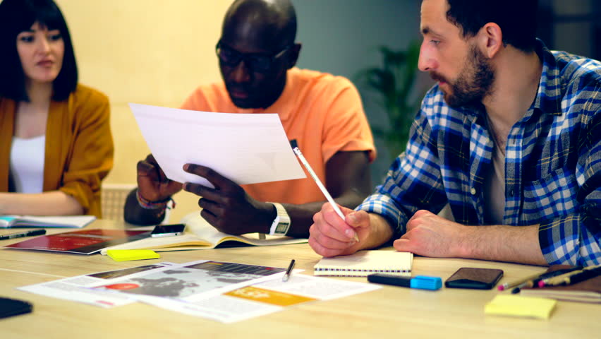 African american male coach checking report and explaining working task to team of young professionals.Multicultural group of male and female colleagues discussing business strategy during meeting