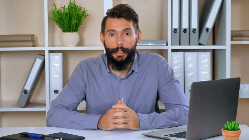 bearded businessman talking with viewers tell about his job. successful emotional employer sitting at the workspace looking at the camera emotionally gesticulates friendly smiling Royalty-Free Stock Footage #1010439104