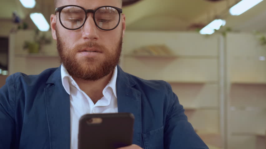 portrait adult businessman using smart phone messaging indoors. handsome man with red beard wearing casual jacket texting message at work or cafe chatting or sharing something or checking email or buy Royalty-Free Stock Footage #1010439854