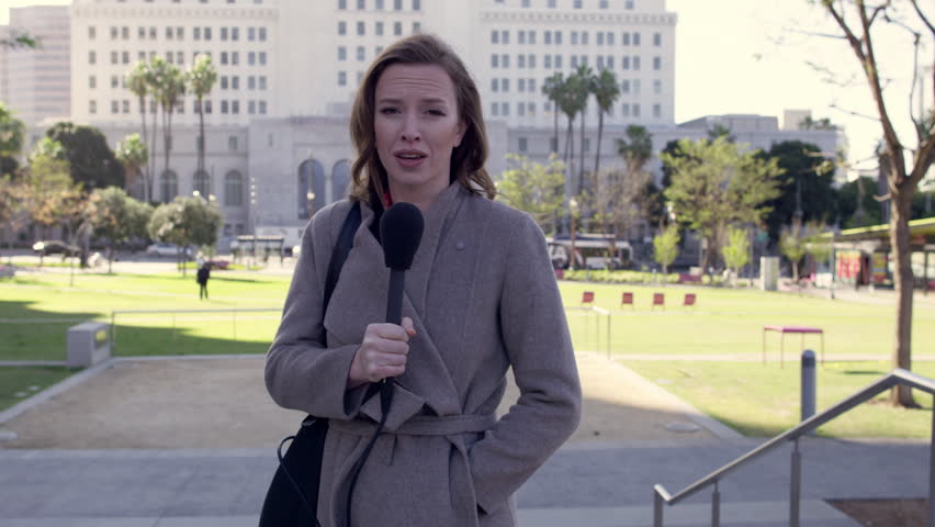 MS smartly dressed female TV reporter talks with microphone with City Hall in background at Grand Park in Downtown Los Angeles. Hand-held, real time 4K UHD. Stabilized, mute