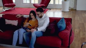 Happy couple in love smiling while watching funny video on modern smartphone using wireless home internet resting on couch. Positive boyfriend together with girlfriend doing shopping online on cellular