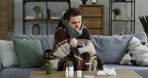 Unwell caucasian man covered with a warm plaid measuring a temperature and having a thermometer in his mouth, shaking as having a fever and a flu. At home.2019-ncov , coronavirus , virus 