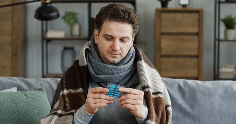Unwell upset man in the warm scarf and plaid sitting on the couch at home, caughing hard and holding pills in a hand. Inside