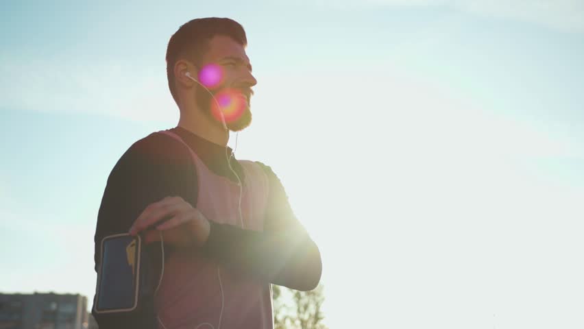 Handsome young guy sportsman doing use phone holder smiling wearing sports clothes running outside listen music sunset stand sport fitness man technology activity training workout