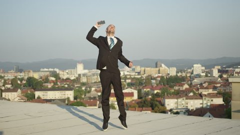 SLOW MOTION CLOSE UP DOF: Genuinely excited businessman jumping for joy when receiving message by mobile phone of closing big deal. Cheerful young man leaping on rooftop punching with hand in air