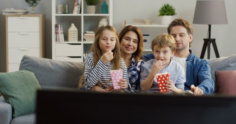 Cheerful caucasian family eating popcorn while sitting on the couch in the living room and watching TV. Portrait shot. Inside