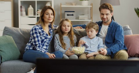 Caucasian young parents with their kids sitting on the couch in the living room, eating popcorn and watching TV. Portrait shot. Indoors
