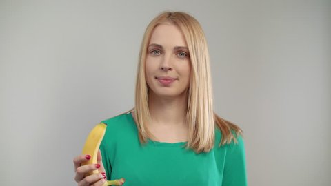 Close of blond woman eating banana at white background. Portrait of blonde girl biting ripe fruit at studio. Vegan woman eating organic food