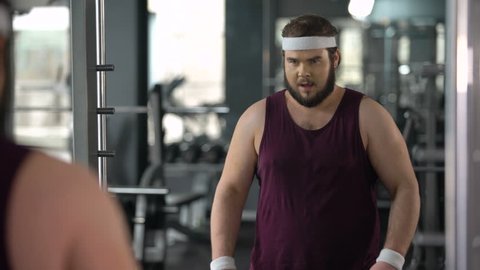 Funny fat man looking at mirror reflection gym and posing, pretending muscular