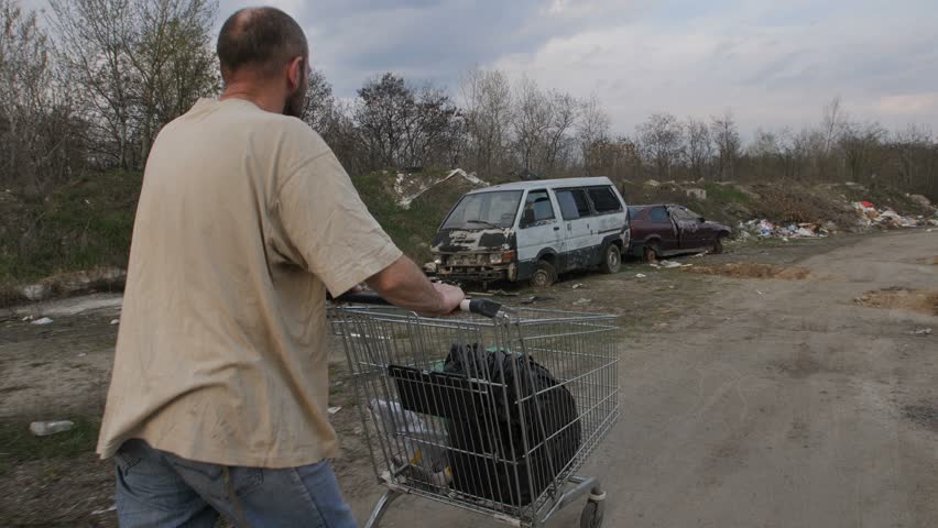 Back view of homeless male walking at landfill site with garbage and old abandoned cars, pushing cart with plastic bags. Homelessness and social issues concept. Steadicam shot. Royalty-Free Stock Footage #1010528120