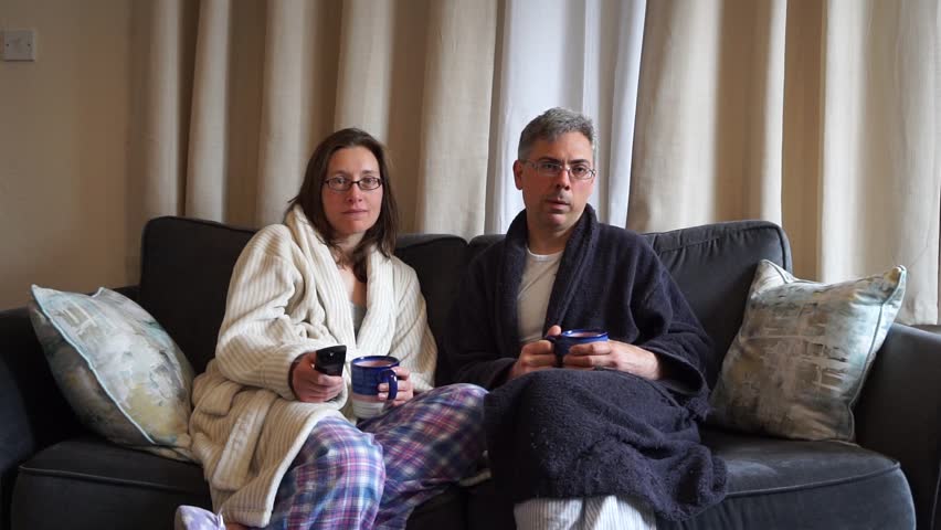 Couple (man and woman) in pajamas are sitting on a sofa channel surfing.  Woman finds a show she wants to watch, but man disagrees.  Man then reluctantly watches the show. Royalty-Free Stock Footage #1010531318