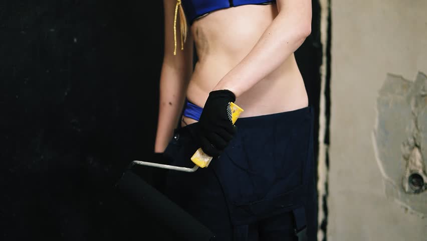 An attractive girl in a bra smears her belly with a black paint roller and continues painting the walls