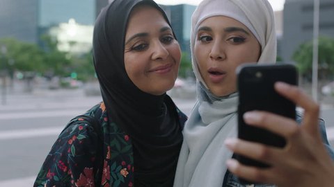 Portrait of young muslim women posing daughter kisses mother on cheek taking selfie photo using smartphone camera technology in urban city background real people series