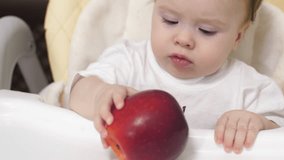Baby is holding red apple in his hands, sitting in children chair.