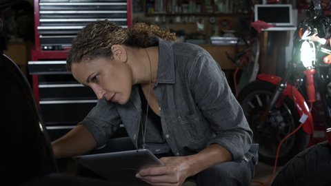 Mature woman referencing mobile tablet and working on motorbike, smiling at camera.