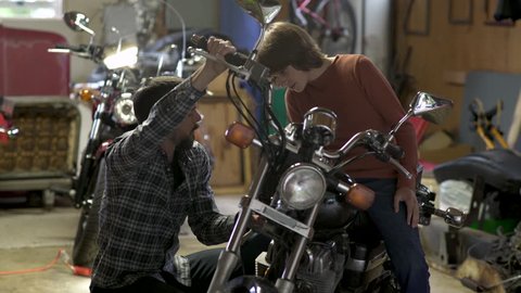 Father mentoring teenage son sitting on motorbike in garage.