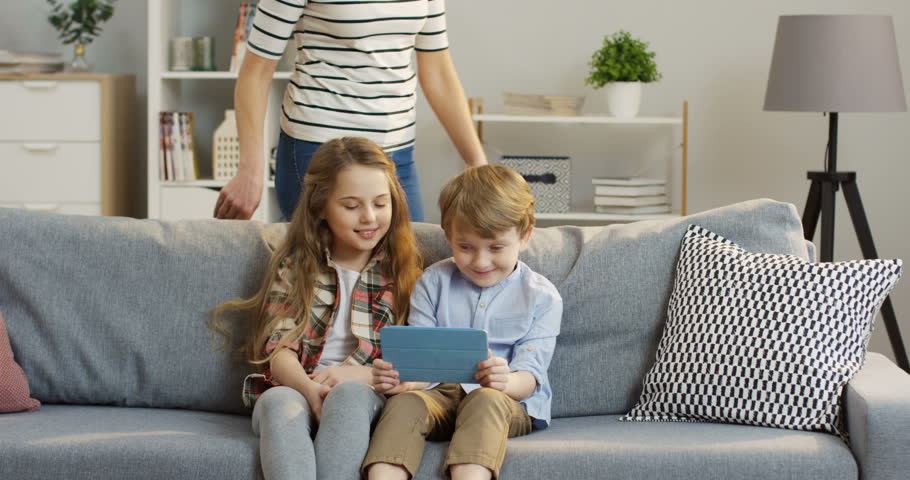 Cute little kids sitting on the couch and watching something on the tablet computer, then their mother coming and hugging them. At home. Inside | Shutterstock HD Video #1010582354