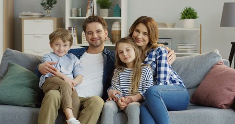 Good looking caucasian family of mother, father, son and daughter sitting on the sofa in the living room and smiling in front of the camera. Portrait shot. Indoors