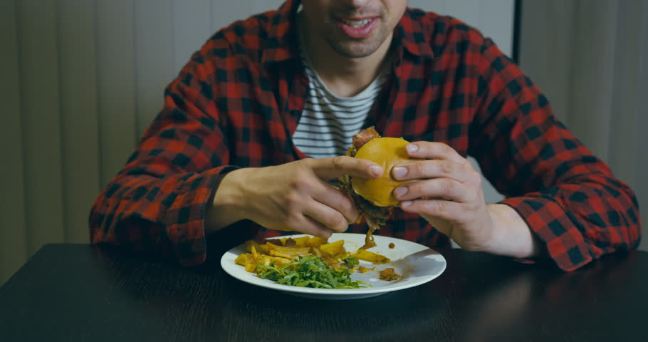A young man in a hipster shirt is eating a hamburger