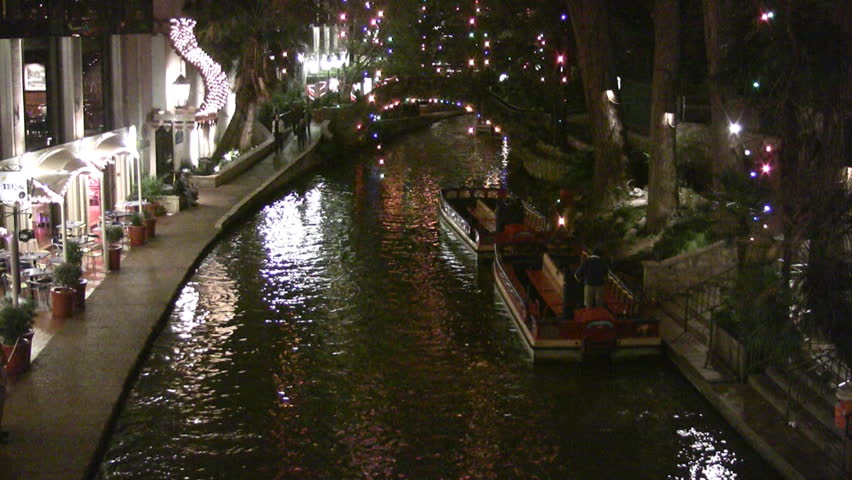 Video night Christmas lights of the San Antonio Texas Riverwalk. Tourists at restaurants and shops. Vacation area. Festive holiday atmosphere. Boats parked on shore. Foot bridge in distance.