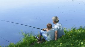 Happy family on summer vacations concept. Father and son fishing together at river bank, man and boy turn to camera and smile happily. Real time full hd video footage.