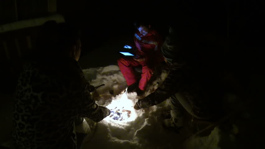 Friends sitting around fire holding sticks with food, checking map on tablet