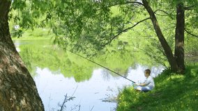 Happy child of 11 years old on spring or summer vacations concept. Boy fishing alone sitting at river grassy bank under huge green tree at beautiful scenic place. Real time full hd video footage.