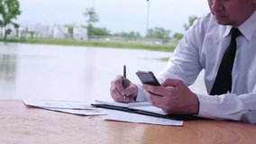 Business man working with mobile phone and document in public park with water pond background - business outdoor working concept