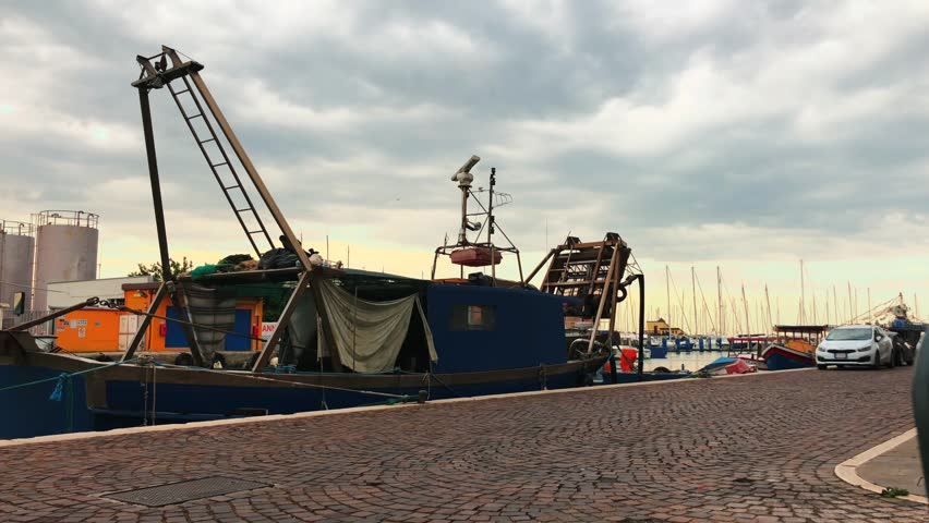 A man cycles down a street next to a canal in an industrial area with a fishing boat in Chioggia Italy near Venice