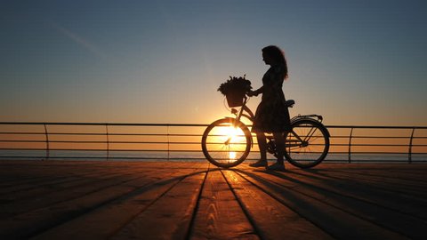 Silhouette of young woman in dress with vintage bicycle and bouquet of flowers walking on wooden embankment near sea during sunrise or sunset. Romantic travel concept. Beautiful scene.