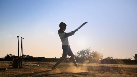 A young boy batting hits the cricket ball for a six in a open field in an Indian village. A slow motion shot of a teenager playing cricket against the sun strikes the cricket ball hard in rural India