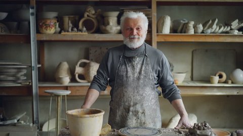 Portrait of senior silver-haired potter in muddy apron standing at table in workshop and looking at camera. Shelves with handmade vases and pots in background.