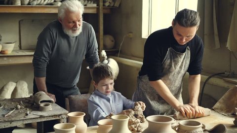 Professional potter is kneading clay on worktable in home studio while his son is helping him and his elderly father watching them from behind. Small family business concept.