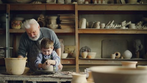 Caring grandfather experienced potter is teaching little boy how to work with clay on potter's wheel. Grandson is making mistake, patient grandpa is helping him.