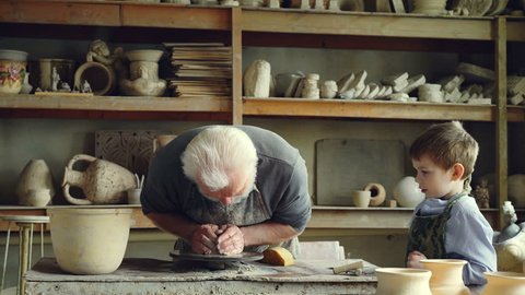 Caring senior grandfather is showing young grandson how to work with clay on throwing-wheel in small workshop. Pottery, family hobby and handicraft concept.