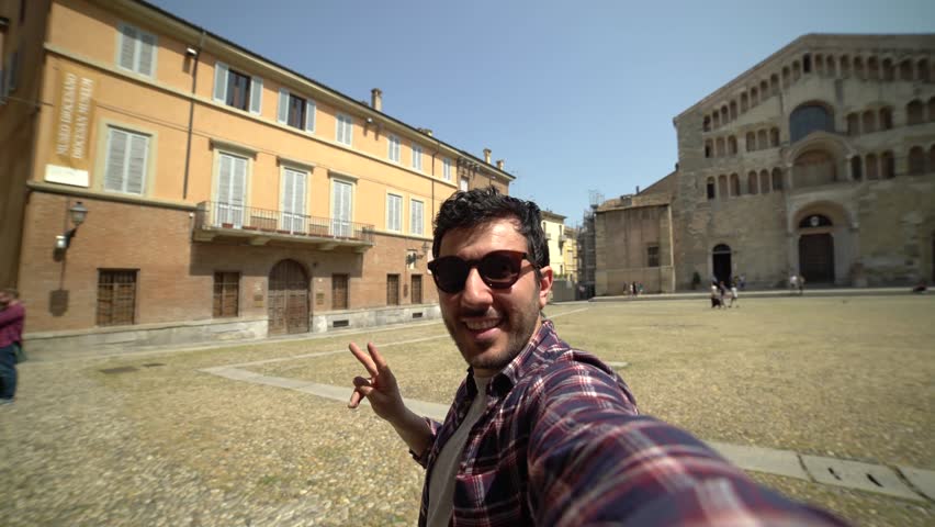 Happytourist takes photo on travel in Parma city. Man backpacker smiling happy taking selfie in the central square with cathedral and famous leaning tower on the background in Parma town.self portrait