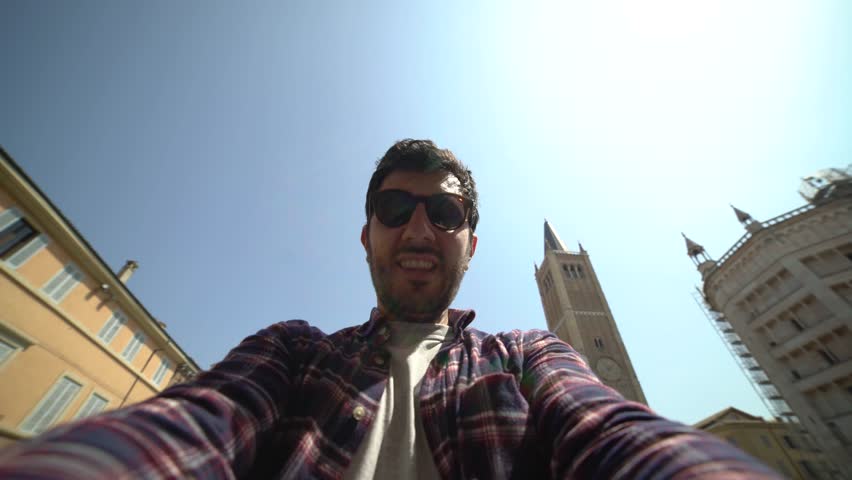 Happytourist takes photo on travel in Parma city. Man backpacker smiling happy taking selfie in the central square with cathedral and famous leaning tower on the background in Parma town.self portrait
