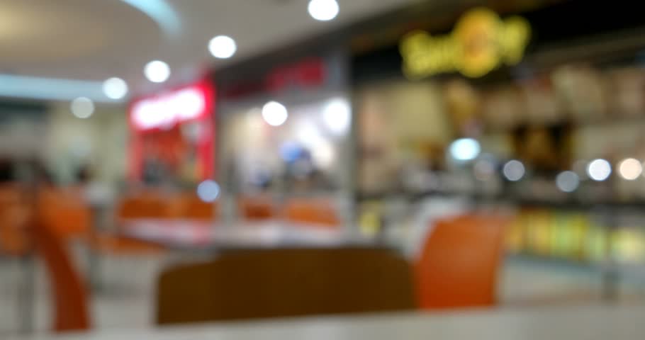 Contemporary dining scene: vibrant food court setting inside bustling mall embodies modern dining experiences, with foreground showcasing hardwood tables and chairs featuring warm textures and subtle sheen that suggest casual atmosphere designed for social interactions. Blurred background reveals lively mix of food stalls with bright neon signage and inviting illumination, familiar fast-food branding emphasizing variety of options available to patrons. Lighting blends soft overhead illumination with bright stall lights, creating dynamic yet cozy environment ideal for showcasing culinary concepts. Color scheme combines warm orange seating with cool gray and cream furnishings, evoking sense of comfort and liveliness perfect for restaurant promotions. This commercial depiction effectively captures communal dining atmosphere and energy of shared public space, making versatile choice for lifestyle blogs and mall advertising. Intimate composition draws viewers into contemporary aesthetic of casual meals and retail environments.