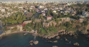 Aerial drone video above water tracking right of rocky cliffs below Kaleici old town to Hidirlik Castle and Karaalioglu Park late afternoon in Antalya, Turkey. 4k at 23.97fps