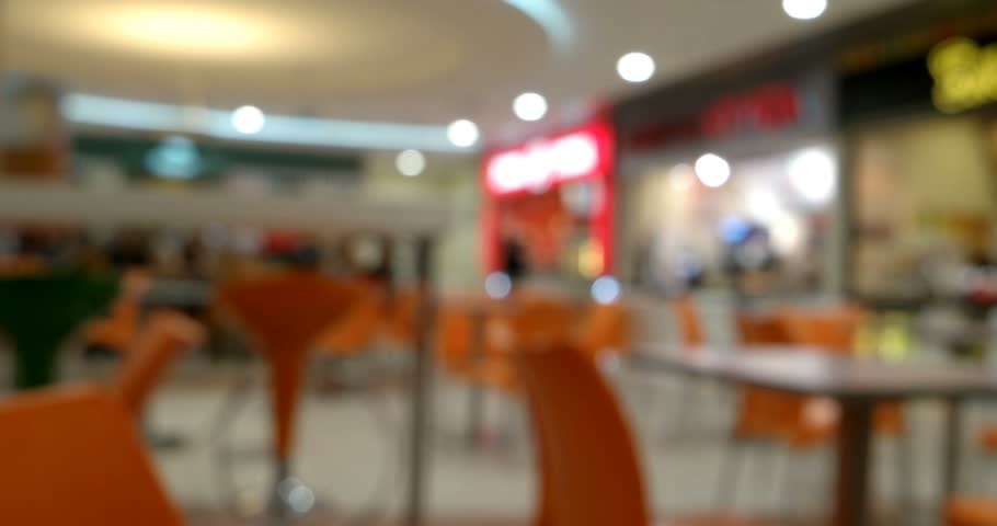 Commercial aesthetic: dynamic walkthrough reveals inviting fast-casual establishment with vibrant contemporary design elements, capturing essence of modern dining culture. Warm ambient illumination from recessed fixtures and accent spotlights bathes expansive food court space in welcoming tones, suggesting late afternoon or early evening hours. Distinctive orange molded plastic seating furniture arranged throughout multiple zones creates visual rhythm, while rich burnt orange upholstered chairs enhance spatial depth from foreground to background. Prominent red neon signage glows in mid-ground, providing striking pop against neutral cream and beige ceiling architecture. Soft focus cinematography with gentle motion conveys relaxed browsing sensation, directing attention toward organized furniture arrangement and architectural elements. Clean, well-maintained environment showcases structured commercial composition ideal for restaurant marketing, hospitality promotions, and interior design portfolios. Color palette dominated by warm oranges, soft creams, accent reds, and whites generates energetic yet comfortable atmosphere appealing to diverse demographics.